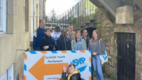 Nine MSYPs are standing outside the SYP Office sign smiling at the camera