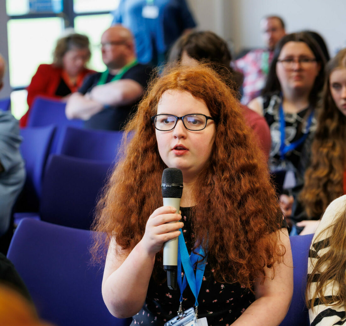Emma Henderson holding a mic in amongst the crowd of MSYPs. She has long red hear and is wearing a black t shirt.