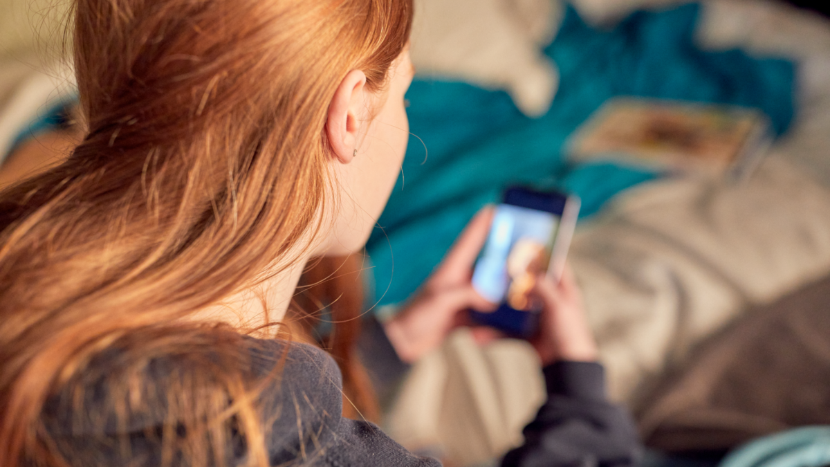Young girl with red hair on her bed using her phone, which is blurred and out of focus.
