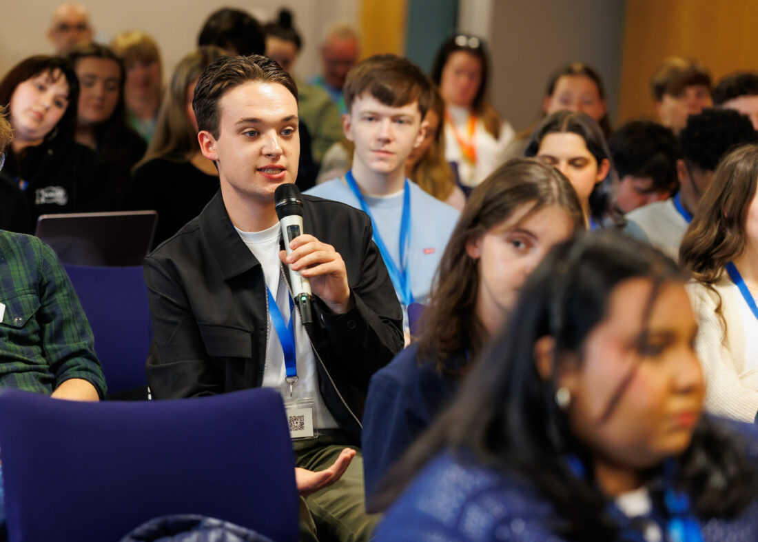 Jack Anderson MSYP wearing a black jacket and a white t-shirt, holding a microphone in the middle of the crowd at the 84rd SYP National Sitting in Stirling.